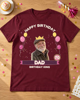 Maroon birthday portrait tee laid on an oak party table with a cupcake, paper horn and confetti, a crown, age 70 badge and Dad lettering.
