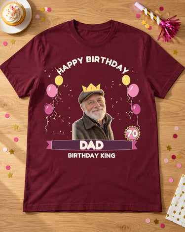 Maroon birthday portrait tee laid on an oak party table with a cupcake, paper horn and confetti, a crown, age 70 badge and Dad lettering.