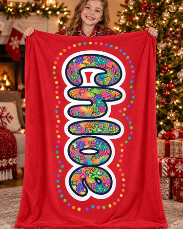 Girl in Christmas pajamas holding a red personalized name blanket reading Chloe next to a decorated Christmas tree