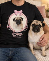 Adult wearer cropped below the chin with a fawn pug in a soft daylight bedroom, wearing the black coquette cameo shirt with the same pug portrait and pink ribbons.