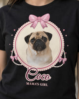 Close-up of a black coquette dog shirt showing the cream oval cameo, scalloped lace, pink satin-style bows, round pearl strands and Coco in outlined script.