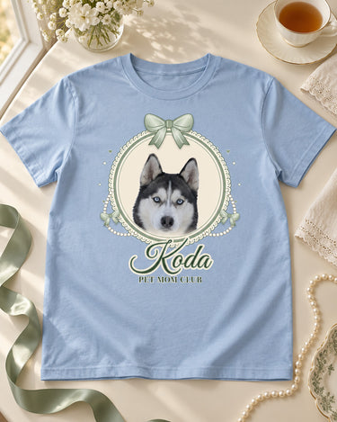 Light blue coquette pet shirt on an ivory tea table with sage ribbon, pearls and a teacup, showing a husky cameo with sage bows and the script name Koda.