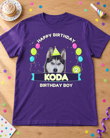 Purple pet birthday tee on an oak table with a cupcake, party horn and confetti, showing a husky in a hat, age 2 and Koda lettering.
