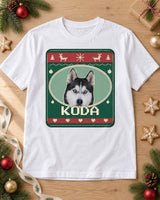 White pet Christmas tee laid on oak with pine branches, ornaments and cookies, showing Koda in an Evergreen Nordic sweater print