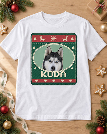 White pet Christmas tee laid on oak with pine branches, ornaments and cookies, showing Koda in an Evergreen Nordic sweater print
