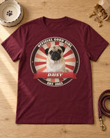 Maroon pet vintage badge t-shirt laid flat on a wooden table with a dog bowl, treats and a leash, a pug in a cherry red sunburst badge with the name Daisy