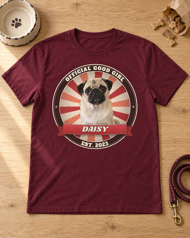 Maroon pet vintage badge t-shirt laid flat on a wooden table with a dog bowl, treats and a leash, a pug in a cherry red sunburst badge with the name Daisy