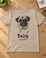 Sand beige pet sketch t-shirt laid flat on a wooden table with pencils, an eraser and a sketchbook, a pug portrait with the name Daisy