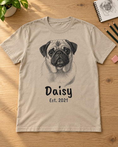 Sand beige pet sketch t-shirt laid flat on a wooden table with pencils, an eraser and a sketchbook, a pug portrait with the name Daisy