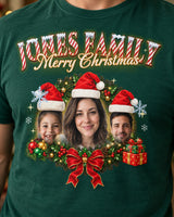 Close-up of the full-color print on a forest green face Christmas t-shirt: three faces in Santa hats in a holly wreath with a red bow