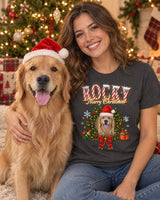 Smiling woman with her golden retriever in a Santa hat beside a Christmas tree, wearing a charcoal pet Christmas t-shirt that shows the same dog in a holly wreath