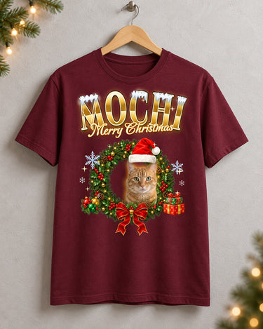 Maroon custom pet Christmas t-shirt on a wooden hanger with the name Mochi in gold lettering and a tabby cat in a Santa hat inside a holly wreath