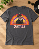 Charcoal retro sunset photo shirt laid on oak beside sunglasses, a vinyl record and a plant, with one capped portrait and JAMES below warm rainbow arcs.
