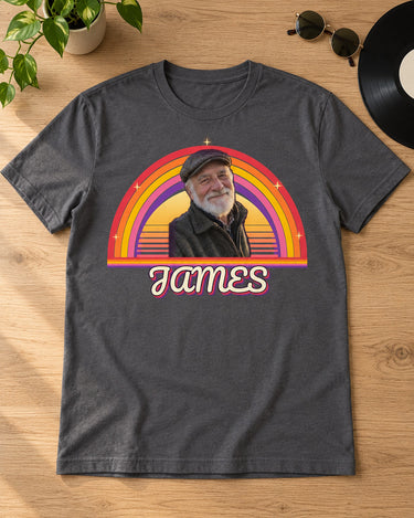 Charcoal retro sunset photo shirt laid on oak beside sunglasses, a vinyl record and a plant, with one capped portrait and JAMES below warm rainbow arcs.