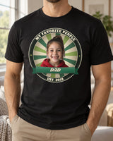 Man wearing a black Dad Est. photo badge t-shirt with a girl's face, forest badge colors and the name Dad
