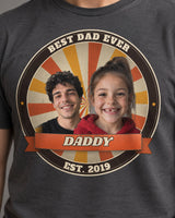 Close-up of the print on a charcoal Dad Est. photo badge t-shirt: two kids' faces in a retro sunburst badge with a ribbon reading Daddy and Est. 2019