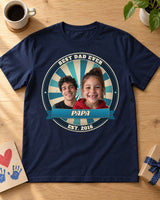 Navy Dad Est. photo badge t-shirt laid flat on an oak table with a Father's Day card, coffee mug and gift, two kids' faces in an ocean badge with the name Papa