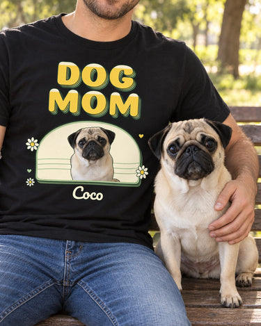 Adult in blue jeans sitting with a fawn pug on a park bench, wearing a black DOG MOM photo tee with the same pug inside a yellow and green daisy arch.