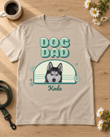Sand DOG DAD photo shirt laid flat on a pale oak table beside a coffee mug, daisies and a leash, showing Koda the husky in a mint and teal arch.