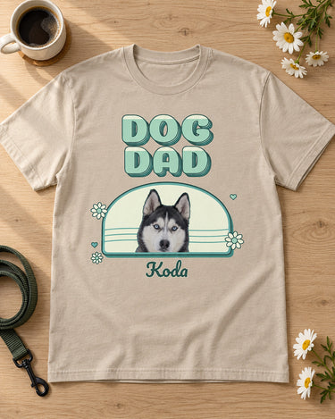 Sand DOG DAD photo shirt laid flat on a pale oak table beside a coffee mug, daisies and a leash, showing Koda the husky in a mint and teal arch.