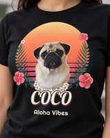 Close view of the black Aloha tee print, with Coco the pug, small cream lei flowers, coral hibiscus, curved sunset wave gaps and outlined script.
