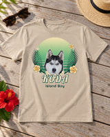 Sand tropical pet photo T-shirt laid on a wooden patio table beside sunglasses, a straw hat and hibiscus, showing Koda in Jungle Green with a lei.