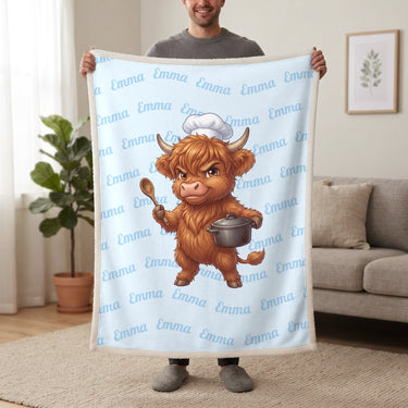 Personalized Highland cow chef blanket in baby blue held up in a living room, with the name Emma repeated in soft waves
