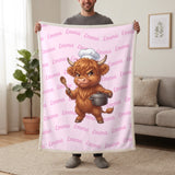 Personalized Highland cow chef blanket in cotton pink held up in a living room, with the name Emma repeated in soft waves