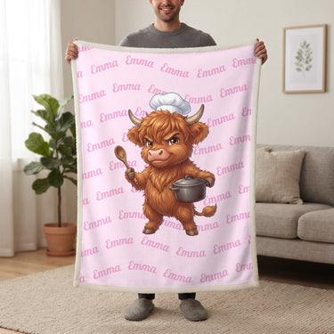 Personalized Highland cow chef blanket in cotton pink held up in a living room, with the name Emma repeated in soft waves