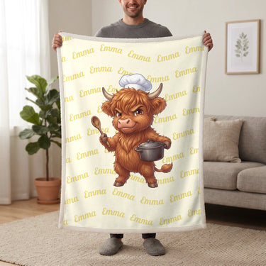 Personalized Highland cow chef blanket in lemon cream held up in a living room, with the name Emma repeated in soft waves
