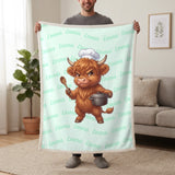 Personalized Highland cow chef blanket in mint held up in a living room, with the name Emma repeated in soft waves