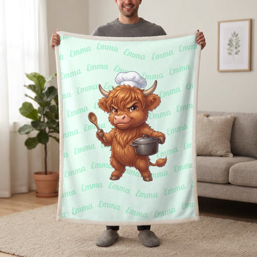 Personalized Highland cow chef blanket in mint held up in a living room, with the name Emma repeated in soft waves