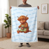 Personalized Highland cow gardener blanket in baby blue held up in a living room, with the name Emma repeated in soft waves
