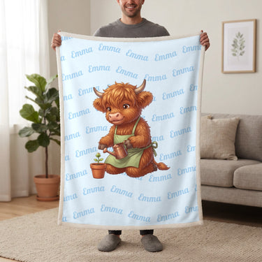 Personalized Highland cow gardener blanket in baby blue held up in a living room, with the name Emma repeated in soft waves
