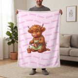 Personalized Highland cow gardener blanket in cotton pink held up in a living room, with the name Emma repeated in soft waves