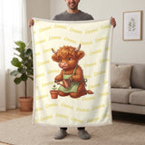 Personalized Highland cow gardener blanket in lemon cream held up in a living room, with the name Emma repeated in soft waves