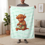 Personalized Highland cow gardener blanket in mint held up in a living room, with the name Emma repeated in soft waves