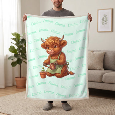 Personalized Highland cow gardener blanket in mint held up in a living room, with the name Emma repeated in soft waves