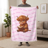 Personalized Highland cow reading blanket in cotton pink held up in a living room, with the name Emma repeated in soft waves