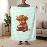 Personalized Highland cow reading blanket in mint held up in a living room, with the name Emma repeated in soft waves