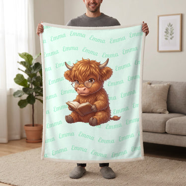 Personalized Highland cow reading blanket in mint held up in a living room, with the name Emma repeated in soft waves