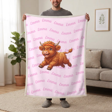 Personalized Highland cow running blanket in cotton pink held up in a living room, with the name Emma repeated in soft waves