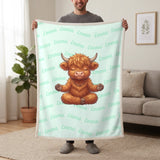 Personalized Highland cow yoga blanket in mint held up in a living room, with the name Emma repeated in soft waves