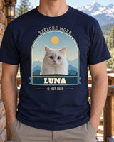 Navy pet adventure shirt on a cabin porch, showing Luna the white cat against blue mountain layers, with Explore More curved above the arch.