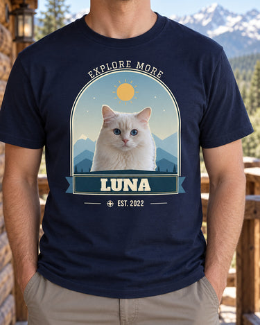 Navy pet adventure shirt on a cabin porch, showing Luna the white cat against blue mountain layers, with Explore More curved above the arch.