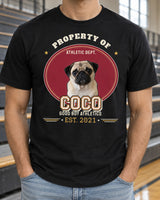 Black Property Of pet varsity shirt beside gym bleachers, with a pug portrait, cream COCO block lettering, gold accents and an EST. 2021 year row.