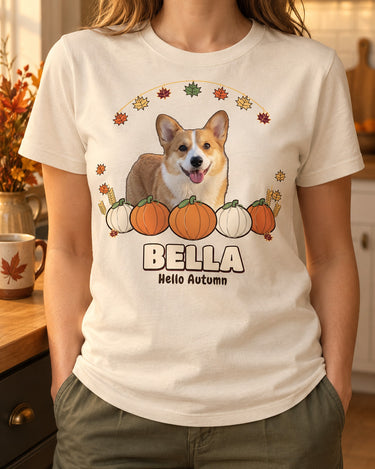 Natural cream personalized fall pet shirt worn in a warm kitchen, with the name BELLA, a dog portrait, orange pumpkins and a hanging autumn leaf garland.