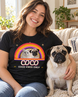 Smiling woman with her fawn pug on a sofa, wearing a black pet retro sunset t-shirt that shows the same pug in front of a 70s rainbow sunset