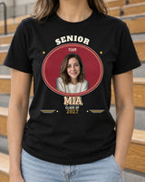 Black senior varsity photo shirt beside gym bleachers, with a portrait in an oval patch, cream MIA lettering and Class of 2027 below.