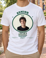 White ALEX senior night photo shirt on a campus walkway, with a portrait in a green and white oval patch, block lettering and the year 2027.